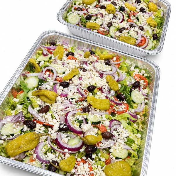 Two catering trays filled with fresh Greek salad, topped with feta, olives, and pepperoncini.