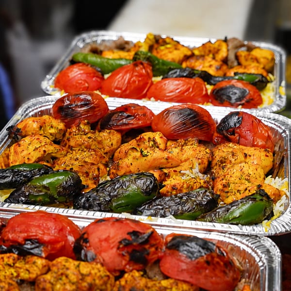 Multiple foil trays filled with grilled chicken, charred tomatoes, and green peppers over rice.