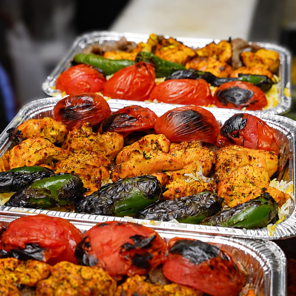 Multiple foil trays filled with grilled chicken, charred tomatoes, and green peppers over rice.